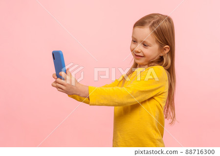 Portrait of very beautiful little girl with freckles and ginger hair smiling into smartphone, taking selfie or communicating with parents on video call. indoor studio shot isolated on pink background 88716300