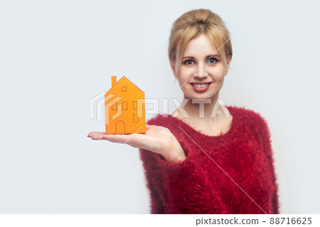 Happy satisfied young realtor woman in red blouse standing, holding paper small house and propose you to planning new home to buy. Indoor, isolated, studio shot, gray background Happy satisfied young realtor woman in red blouse standing, holding paper small house and propose you to planning new home to buy. Indoor, isolated, studio shot, gray background 88716625