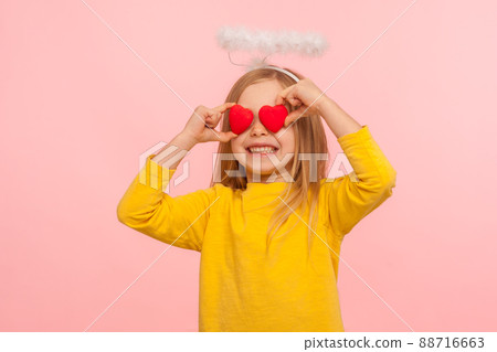 Portrait of nice angelic happy little girl with halo over head covering eyes with toy hearts and smiling sincerely, metaphor of look full of love. indoor studio shot isolated on pink background 88716663