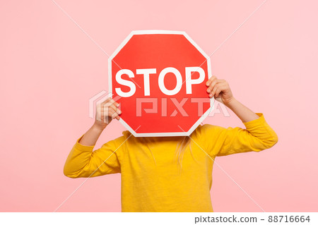 Unrecognizable child in yellow sweatshirt covering face with Stop symbol, anonymous person holding red traffic sign, warning about road safety rules. indoor studio shot isolated on blue background 88716664