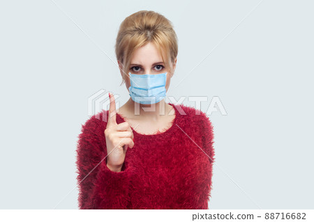Portrait of serious young woman with surgical medical mask in red blouse standing and looking at camera with warning finger and alarming about health care. indoor shot isolated on gray background. 88716682