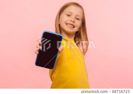 Happy smiling preschool girl holding cellphone to camera with joyful enthusiastic expression, advertising mobile device or children's application, focus on smartphone. indoor studio shot, isolated 88716725