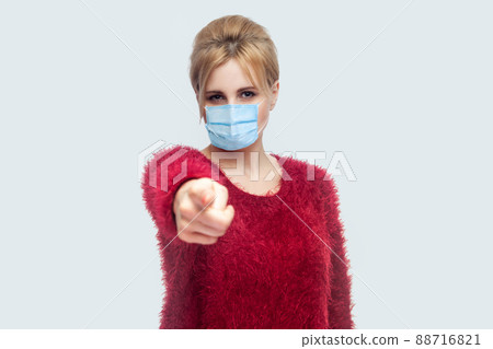 Portrait of serious woman with surgical medical mask in red blouse standing, pointing and looking at camera and blame. indoor studio shot isolated on gray background. 88716821