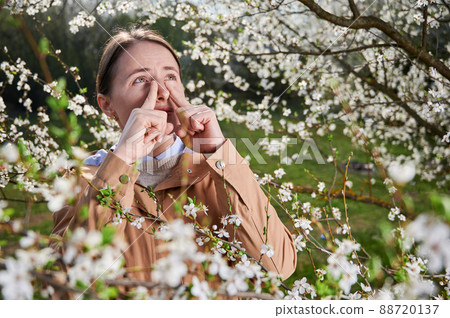 Woman allergic suffering from seasonal allergy at spring, posing in blossoming garden at springtime. Young woman sneezing in front of blooming tree. Spring allergy concept Woman allergic suffering from seasonal allergy at spring, posing in blossoming garden at springtime. Young woman sneezing in front of blooming tree. Spring allergy concept 88720137