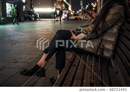Beautiful elegant women's legs in black glossy loafers. A woman sits on a bench on a night street in the city in autumn 88721491