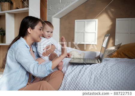 Modern young mother holding her baby daughter greeting her friend or husband on video call on laptop Modern young mother holding her baby daughter greeting her friend or husband on video call on laptop 88722753