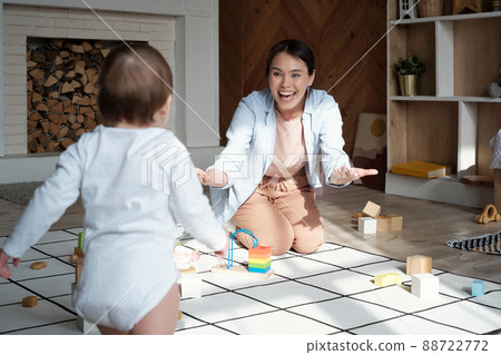 Cheerful young mother sitting on floor in living room encouraging her baby to walk towards her 88722772
