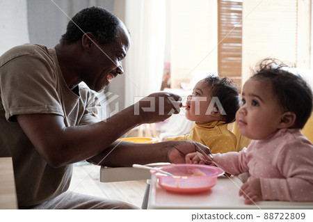 Cheerful mature African American father taking care of his baby daughters feeding them with fruit puree Cheerful mature African American father taking care of his baby daughters feeding them with fruit puree 88722809