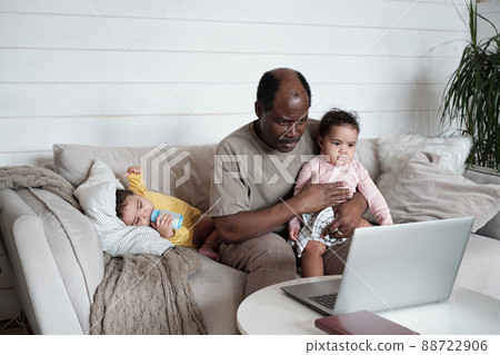Portrait of busy African American dad sitting on sofa in living room taking care of his twin babies and working in Internet 88722906