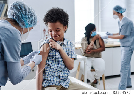 African brave boy getting vaccination in shoulder from nurse during procedure at hospital with other people in background 88724572