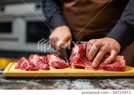 A butcher in an apron in the kitchen cuts pork on a wooden board. Steak. A butcher in an apron in the kitchen cuts pork on a wooden board. Steak. 88724931