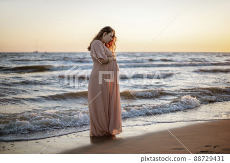 Young pregnant woman with a beautiful sea view on the background. Happy and calm pregnant woman with long hair and pink dress standig on the beach. Romantic view, ocean, sunset, maternity. Young pregnant woman with a beautiful sea view on the background. Happy and calm pregnant woman with long hair and pink dress standig on the beach. Romantic view, ocean, sunset, maternity. 88729431