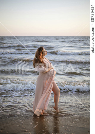 Young pregnant woman with a beautiful sea view on the background. Happy and calm pregnant woman with long hair and pink dress standig on the beach. Romantic view, ocean, sunset, maternity. Young pregnant woman with a beautiful sea view on the background. Happy and calm pregnant woman with long hair and pink dress standig on the beach. Romantic view, ocean, sunset, maternity. 88729434