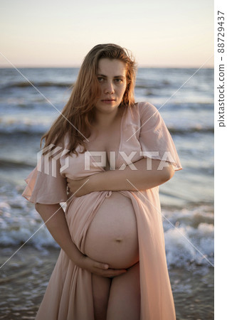 Young pregnant woman with a beautiful sea view on the background. Happy and calm pregnant woman with long hair and pink dress standig on the beach. Romantic view, ocean, sunset, maternity. Young pregnant woman with a beautiful sea view on the background. Happy and calm pregnant woman with long hair and pink dress standig on the beach. Romantic view, ocean, sunset, maternity. 88729437