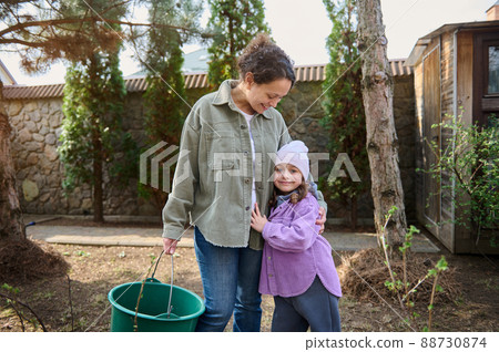 Happy baby girl hugging her loving mother while watering plants in the backyard of their country house, standing together in the garden and enjoying early spring gardening Happy baby girl hugging her loving mother while watering plants in the backyard of their country house, standing together in the garden and enjoying early spring gardening 88730874
