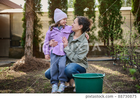 Happy mother and daughter enjoying gardening in the backyard of their country house, watering baby plants in the looseness ground Happy mother and daughter enjoying gardening in the backyard of their country house, watering baby plants in the looseness ground 88730875