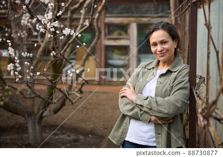 Confident middle aged Caucasian woman gardener enjoying an early spring day standing in her garden plot against the blossoming apricot fruit trees background Confident middle aged Caucasian woman gardener enjoying an early spring day standing in her garden plot against the blossoming apricot fruit trees background 88730877