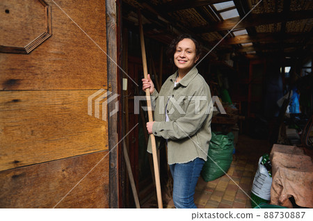 Happy female gardener farmer with gardening tools standing in her wooden rural workshop, enjoying gardening on a beautiful early spring day Happy female gardener farmer with gardening tools standing in her wooden rural workshop, enjoying gardening on a beautiful early spring day 88730887