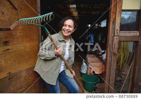 Happy pleasant mature female gardener holding a rake and smiling with a cheerful toothy smile while standing in her wooden rural workshop with gardening tools enjoying home gardening on a spring day Happy pleasant mature female gardener holding a rake and smiling with a cheerful toothy smile while standing in her wooden rural workshop with gardening tools enjoying home gardening on a spring day 88730889
