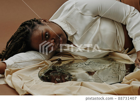 Portrait of African young woman lying near the mirror with water drops on surface and looking at camera Portrait of African young woman lying near the mirror with water drops on surface and looking at camera 88731407