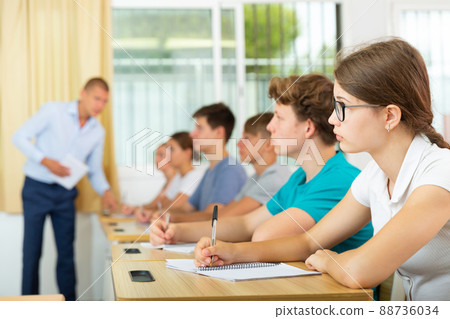 Teen girl in glasses listening to lecturer and writing in notebook in classroom Teen girl in glasses listening to lecturer and writing in notebook in classroom 88736034