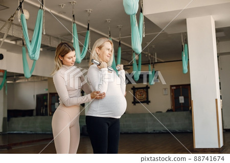 Pregnant woman doing yoga with personal trainer. Yoga trainer assisting pregnant woman while doing exercises with a fitball and dumbbells. Blonde pregnant woman wearing white shirt. 88741674