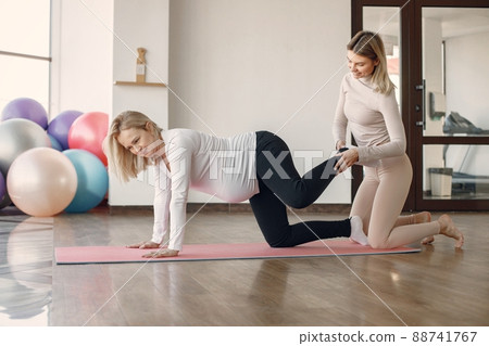 Pregnant woman doing yoga with personal trainer. Yoga trainer assisting pregnant woman while doing exercises. Blonde pregnant woman wearing white clothes. 88741767