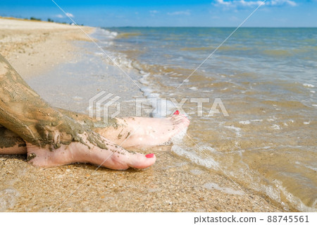 View of beautiful female feet with bright red pedicure on the sand of the beach. The ankles of the feet are smeared with healing mud. Skin rejuvenation while relaxing at sea. Spa. Copy space View of beautiful female feet with bright red pedicure on the sand of the beach. The ankles of the feet are smeared with healing mud. Skin rejuvenation while relaxing at sea. Spa. Copy space 88745561
