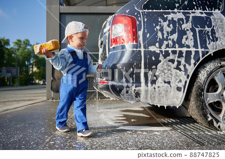 Kid boy studying work at car wash. Toddler child, holding yellow foam sponge in his right hand, examining soap car and preparing to start cleaning the back part of transport. Kid boy studying work at car wash. Toddler child, holding yellow foam sponge in his right hand, examining soap car and preparing to start cleaning the back part of transport. 88747825
