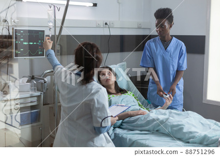 Doctor setting up central line catether with iv drip medication to patient while nurse is putting pulse oximeter on finger. Woman in hospital bed looking at doctor giving her intravenous antibiotics. Doctor setting up central line catether with iv drip medication to patient while nurse is putting pulse oximeter on finger. Woman in hospital bed looking at doctor giving her intravenous antibiotics. 88751296