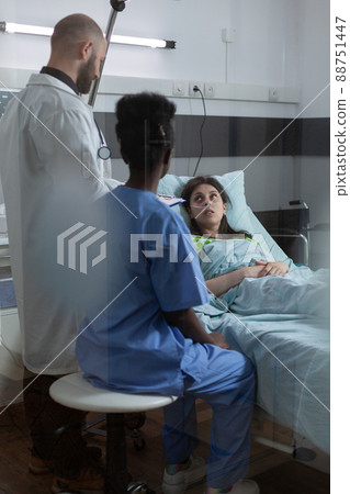 Through the window view of doctor with clipoboard and nurse in private ward consulting recovering patient after medical procedure. Patient with nasal cannula looking at health care professionals. Through the window view of doctor with clipoboard and nurse in private ward consulting recovering patient after medical procedure. Patient with nasal cannula looking at health care professionals. 88751447