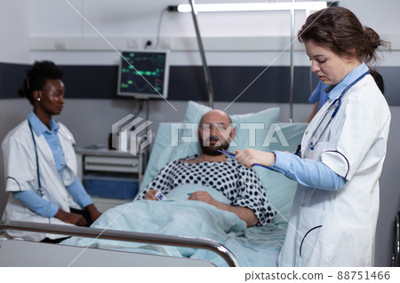 Middle aged man recovering lying in hospital bed attended by team of doctors after surgical intervention. Doctor with stethoscope reading medical chart while nurse is monitoring patient vitals. Middle aged man recovering lying in hospital bed attended by team of doctors after surgical intervention. Doctor with stethoscope reading medical chart while nurse is monitoring patient vitals. 88751466