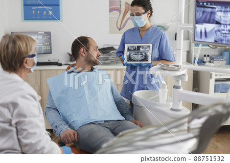 Dentist assistant with face mask holding tabet computer explaining teeth radiography to sick patient during dentistry consultation in stomatological office room. Team working at caries treatment 88751532