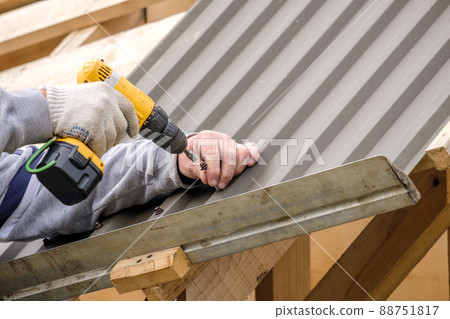 Male hands in gloves with a screwdriver screw the roofing sheet to the roof. Male hands in gloves with a screwdriver screw the roofing sheet to the roof. 88751817