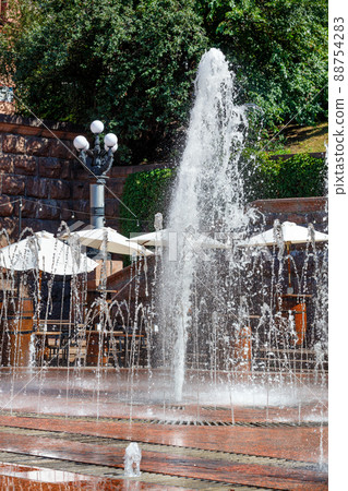City fountain against the background of a summer cafe in a sunny park. City fountain against the background of a summer cafe in a sunny park. 88754283