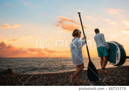 Summer vacations. Father and daughter are walking along the beach with a sup board. Sea and the sunset in the background. Back view. Sea surfing Summer vacations. Father and daughter are walking along the beach with a sup board. Sea and the sunset in the background. Back view. Sea surfing 88755578