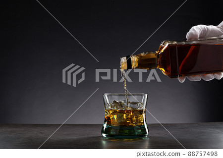 White gloved hand pouring whisky into a glass with ice on dark background, with copy space. White gloved hand pouring whisky into a glass with ice on dark background, with copy space. 88757498