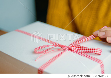 Girl's fingers hold red checkered ribbon on gift box. Holiday mood. Festive surprise for Christmas, New Year or Birthday. 88760527