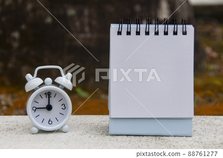 Alarm clock pointing at 9 o'clock with blank notepad. Blurred park background. Copy space concept 88761277