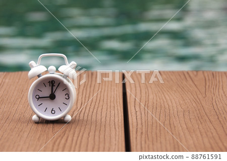 Alarm clock on wooden floor with blurred swimming pool background. The clock set at 9 o'clock. Morning concept. 88761591
