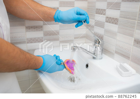 The dentist rinses the dental impression with water under pressure. Close-up of dentist's hands in blue gloves The dentist rinses the dental impression with water under pressure. Close-up of dentist's hands in blue gloves 88762245