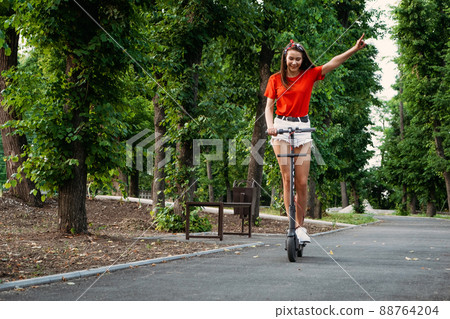E-bikes, scooters safety. Young woman riding e scooter in urban environment. Trendy woman on electric scooter in city park E-bikes, scooters safety. Young woman riding e scooter in urban environment. Trendy woman on electric scooter in city park 88764204