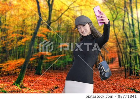 一個微笑的女孩在色彩絢麗的秋季森林中散步，在大自然的背景下用粉色智能手機自拍。 88771841