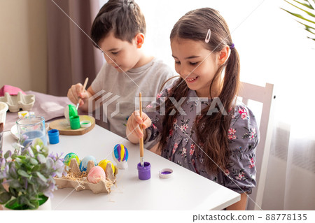 A smiling little girl and boy sits at a white table near the window, paints eggs 88778135