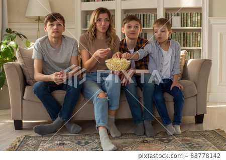 Happy family, a woman and three boys, are sitting on the sofa in the living room with a bowl of popcorn, watching TV 88778142