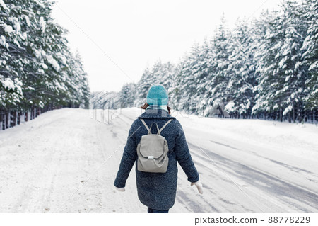 a girl in a gray coat with a gray backpack walking the side of a winter road, next to a winter forest of pine trees 88778229