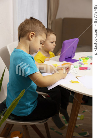 Happy kids with yellow and blue shirts doing arts and crafts together at their desk and watching cartoon on digital tablet. Children have fun. Boys play at home. 88778488