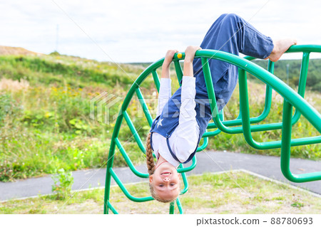 Caucasian cheerful girl in a Blue summer overalls hanging upside down on the Green horizontal bar playground, looking at camera, smiling. School change, physical training. Happiness, childhood 88780693