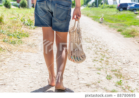 The girl goes to the store barefoot along the village road, bare feet, rear view. Eco-friendly reusable mesh bag, handmade, macrame. Eco-friendly concept without waste. The girl goes to the store barefoot along the village road, bare feet, rear view. Eco-friendly reusable mesh bag, handmade, macrame. Eco-friendly concept without waste. 88780695