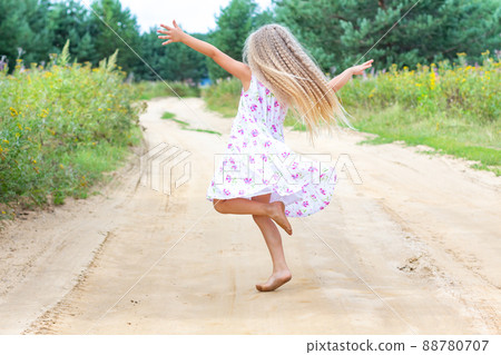 A girl with long blond curly hair is dancing, spinning on a forest road. Childhood, happiness, freedom of movement, dancing, reuniting with nature. Beautiful summer landscape. 88780707